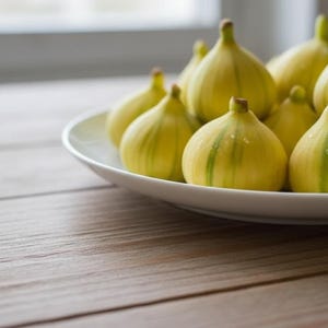 May include: A white ceramic plate filled with ripe figs. The figs are yellow with green stripes and are arranged on a wooden surface. The background is blurred, with a window visible.