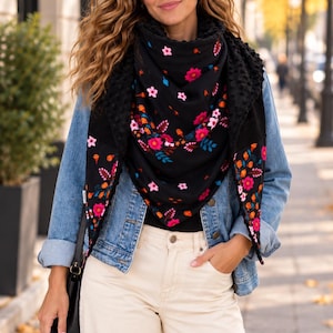 May include: A black triangular scarf featuring a vibrant floral pattern in shades of pink, orange, and blue. The scarf has a textured, bumpy fabric on the reverse side and is worn over a blue denim jacket and cream-coloured trousers.