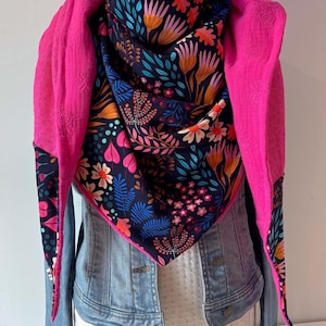 May include: A triangular scarf featuring a dark navy blue base with a vibrant floral and botanical print in shades of pink, orange, and light blue. The scarf is lined with a bright fuchsia pink fabric and is draped over a light blue denim jacket.