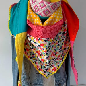 Peut inclure: Un foulard triangulaire en patchwork composé d'un mélange vibrant de tissus, incluant des panneaux unis jaune, bleu sarcelle et rouge, un motif à pois blancs, un imprimé floral rose, un motif géométrique en losanges et des formes abstraites.