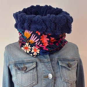 Peut inclure: Un tour de cou doublé de polaire bleu marine avec un motif floral. Le tour de cou présente une doublure en polaire duveteuse bleu foncé et un extérieur à motifs avec des fleurs roses, oranges et blanches sur fond marine. Il est présenté sur une veste en jean.