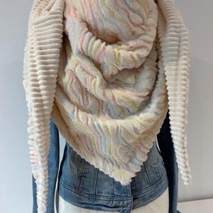 May include: A cream-coloured, hooded scarf with a soft, textured appearance. The scarf features a ribbed design on the outer edges and a wavy, pastel-coloured pattern on the inner layer. It is draped over a denim jacket.