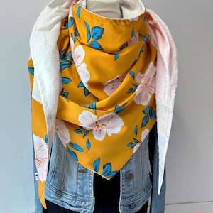 Peut inclure: Écharpe jaune moutarde avec un motif floral de fleurs blanches et roses et de feuilles bleues. L'écharpe est superposée à un tissu blanc texturé. Une veste en jean et un haut noir sont visibles.