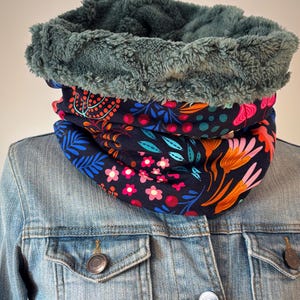 May include: A cozy neck warmer with a dark teal faux fur lining and a floral patterned exterior. The pattern features pink, orange, and blue flowers and leaves on a navy blue background. The neck warmer is shown on a denim jacket.