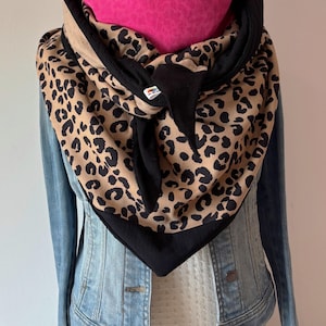 Peut inclure: Écharpe élégante avec un imprimé léopard beige et noir, bordée de noir. Elle est superposée à un chauffe-cou rose vif. L'écharpe est portée sur une veste en jean.