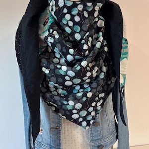 Peut inclure: Écharpe noire ornée d'un motif botanique avec des feuilles bleu-vert et des accents blancs. Elle est drapée sur une veste en jean et un haut blanc, créant un look superposé. Le tissu de l'écharpe a un aspect doux et fluide.