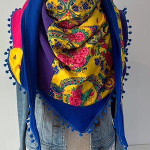 Peut inclure: Un foulard triangulaire en tissu jaune vif avec un motif floral rose et vert, superposé à des sections de tissu violet et bleu uni. Les bords sont ornés de petits pompons bleus, et le foulard est drapé sur une veste en jean bleu clair.