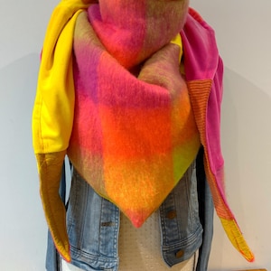 Peut inclure: Une écharpe vibrante et colorée avec un motif géométrique dans les tons de rose, orange et jaune. L'écharpe est drapée sur une veste en jean, mettant en valeur son design audacieux et sa texture. Les extrémités de l'écharpe sont jaunes et roses.