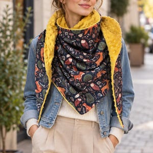 May include: A triangular scarf featuring a dark navy blue fabric with a repeating pattern of orange foxes, brown owls, deer, and green foliage. The reverse side is a solid mustard yellow textured fabric with a raised, dimpled dot pattern.