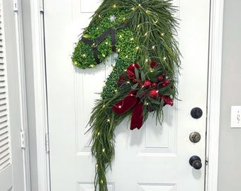 May include: A horse-shaped Christmas wreath made of green foliage, adorned with small white lights and a red bow. The wreath is hanging on a white door, creating a festive holiday decoration.