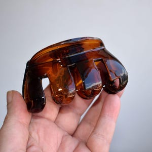 Può includere: Un fermaglio per capelli a forma di artiglio, di colore marrone traslucido con finitura lucida e bordo superiore arrotondato. L'accessorio presenta diversi denti a incastro progettati per fissare i capelli, con un classico motivo tartarugato.