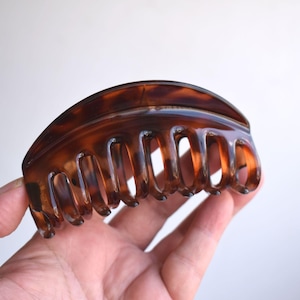Könnte beinhalten: Eine gebogene Haarspange mit einem glänzenden, durchscheinenden braun-schwarzen Schildpattmuster. Das Accessoire hat eine halbkreisförmige Oberkante und eine Reihe von ineinandergreifenden Zähnen an der Unterseite, um das Haar zu fixieren.