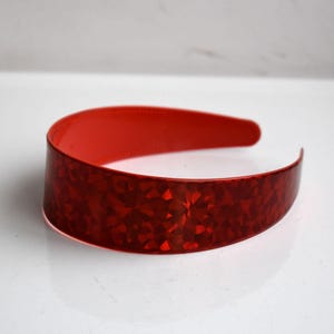 Può includere: Una fascia per capelli rossa con un motivo geometrico strutturato. La fascia è di un colore rosso vibrante e ha una superficie lucida e riflettente. Questo accessorio per capelli è un'aggiunta elegante a qualsiasi outfit.