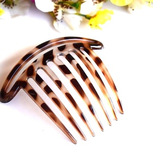 May include: A tortoiseshell hair comb with a curved top and six tines. The comb has a pattern of brown spots on a translucent background. The hair accessory is designed for styling and securing hair.