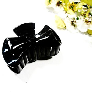 May include: A glossy black hair claw clip. The clip is made of a hard plastic and has a bow-like design. The clip is designed to hold hair in place. The background is white with yellow and white flowers.