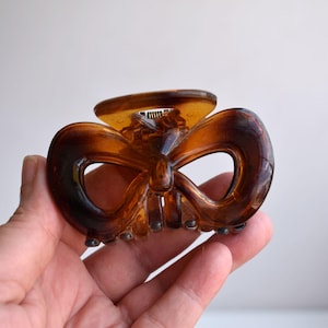 May include: A translucent brown tortoiseshell hair claw clip featuring a bow-shaped design with two symmetrical loops. The accessory has a spring-loaded mechanism with interlocking teeth along the bottom edge to secure hair in place.