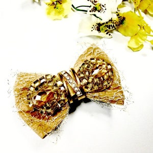 May include: A gold-toned bow with a central embellishment of sparkling crystals and rhinestones. The bow features a textured, layered design with a delicate, net-like trim. Yellow and white flowers are in the background.
