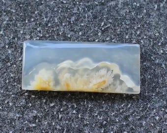 May include: A rectangular, polished gemstone with a cloudy, landscape-like pattern. The stone features a translucent, light blue base with white and yellow cloud-like inclusions. The overall effect is reminiscent of a miniature sky scene.