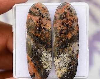 May include: Two oval gemstones with a hole drilled at the top. The stones feature a mix of orange, black, and white, with a speckled pattern. The gemstones are presented in a white box.