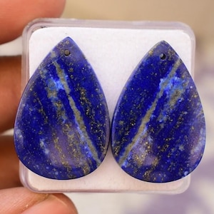 May include: Two teardrop-shaped, polished lapis lazuli stones. The stones are a deep blue with streaks of gold and white. Each stone has a small hole at the top for stringing. The stones are displayed in a clear plastic case.