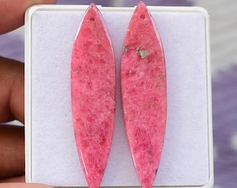 May include: Two elongated, pink gemstone pendants with a hole at the top for stringing. The pendants have a polished, smooth surface and a speckled appearance. The gemstones are displayed in a white box.