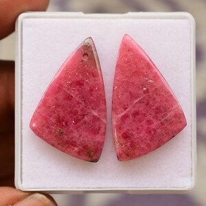 Puede incluir: Dos piedras preciosas triangulares pulidas de color rosa vibrante. Cada piedra tiene un peque&ntilde;o agujero en la parte superior, lo que sugiere que est&aacute;n destinadas a la fabricaci&oacute;n de joyas. Las piedras est&aacute;n sobre un fondo blanco.