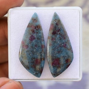 May include: Two teardrop-shaped gemstones with a blue-gray base color and flecks of red and brown. Each stone has a small hole at the top. The stones are displayed in a white plastic case.