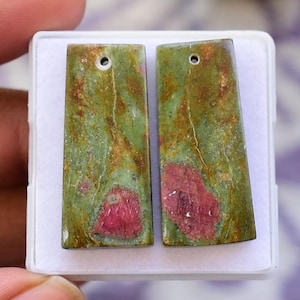 Könnte beinhalten: Zwei rechteckige, polierte Edelsteinanhänger. Die Anhänger sind grün mit roten und braunen Einschlüssen und haben jeweils ein kleines Loch oben. Die Edelsteine werden in einer weißen Box präsentiert.