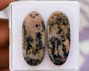 May include: Two oval gemstones with a hole drilled at the top. The stones feature a mix of black, tan, and gold colours. The gemstones are presented in a white plastic case.
