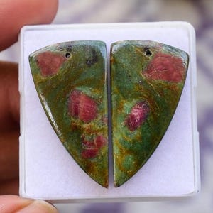 Könnte beinhalten: Zwei polierte Edelsteinanhänger in Dreiecksform. Sie sind grün mit roten und rosa Einschlüssen und haben jeweils ein kleines Loch oben. Die Edelsteine werden in einem weißen Kunststoffetui präsentiert.