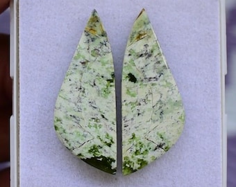 Puede incluir: Dos piedras preciosas en forma de lágrima con un patrón veteado verde claro y blanco, acentuado con vetas verdes oscuras y negras. Las piedras se exhiben en una caja blanca, mostrando su belleza natural y veteado único.