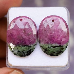 Peut inclure: Deux pierres précieuses ovales avec une surface à facettes. Les pierres sont un mélange de rose, de vert et de noir. Chaque pierre a un petit trou en haut. Les pierres sont présentées dans un récipient en plastique transparent.