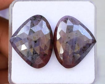 Puede incluir: Dos piedras preciosas facetadas en forma de escudo con un tono púrpura intenso y marrón rojizo. Las piedras presentan una superficie pulida de múltiples facetas con inclusiones oscuras visibles, dispuestas una al lado de la otra sobre una base blanca.