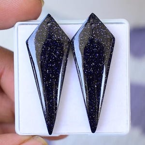 Può includere: Due ciondoli di pietre preziose blu scuro con un aspetto scintillante e maculato. Ogni ciondolo ha una forma a diamante allungata con un piccolo foro nella parte superiore. I ciondoli sono esposti in una scatola bianca.