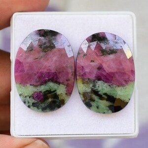 Puede incluir: Dos piedras preciosas ovaladas con corte facetado. Las piedras muestran una mezcla de colores rosa, verde y negro, con una superficie pulida. Las piedras están en una caja blanca, para la creación de joyas.