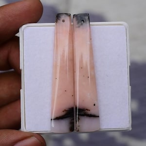 Puede incluir: Dos cuentas colgantes alargadas y cónicas hechas de ópalo rosa pulido con inclusiones dendríticas negras naturales. Cada piedra lisa de reverso plano presenta un pequeño orificio perforado en la parte superior para colgar, mostrando una superficie de tono melocotón.