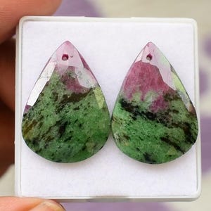 Könnte beinhalten: Zwei tropfenförmige Edelsteine mit einem Loch oben. Die Steine weisen eine Mischung aus grünen und roten Farbtönen auf, mit einer polierten, facettierten Oberfläche. Die Edelsteine sind vor einem weißen Hintergrund platziert.