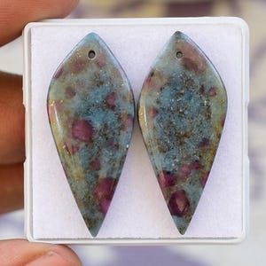 Puede incluir: Dos piedras preciosas en forma de lágrima con un agujero en la parte superior. Las piedras tienen un color base azul-gris con inclusiones rojas y marrones. Están colocadas en un recipiente de plástico blanco.