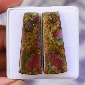 Könnte beinhalten: Zwei rechteckige Edelsteinanhänger mit einem Loch oben zum Auffädeln. Die Anhänger haben ein gesprenkeltes Aussehen mit Grüntönen, Braun und Rot. Die Anhänger werden in einer weißen Box präsentiert.