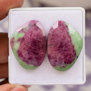 May include: Two gemstone pendants with a faceted cut. The pendants are a mix of deep red and light green colors. Each stone has a hole drilled at the top for stringing. The stones are displayed in a white box.