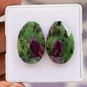 May include: Two oval gemstones with a green base, speckled with dark spots and a magenta section. The gems are set in a white box, highlighting their polished surfaces and natural patterns. These gems are likely for jewellery making.