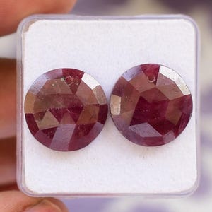 Peut inclure: Deux pierres précieuses rondes et facettées d'un rouge profond. Les gemmes sont présentées dans un boîtier en plastique transparent, mettant en valeur leurs coupes géométriques et leurs surfaces polies. Probablement des rubis.