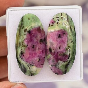 Könnte beinhalten: Zwei ovale Edelsteine mit einer lebendigen Farbmischung. Die Steine weisen eine auffällige Kombination aus tiefem Rosa, Grün und Schwarz auf. Jeder Stein hat ein Loch an der Oberseite, was darauf hindeutet, dass sie für Schmuck bestimmt sind.