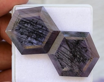 Puede incluir: Dos gemas hexagonales en tonos púrpura oscuro con inclusiones negras. Las gemas se exhiben sobre una superficie blanca, resaltando su diseño multifacético y sus patrones naturales. Las piedras están cortadas con precisión, mostrando sus estructuras internas únicas.
