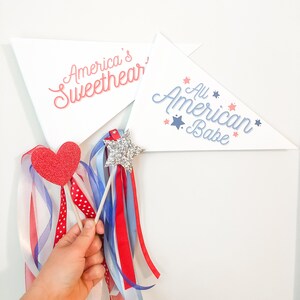 May include: Two white triangle pennants with red, white, and blue stars and text. The pennants say "America's Sweetheart" and "All American Babe".  A red heart-shaped wand with white and blue ribbon streamers and a silver glitter star wand with white and blue ribbon streamers.