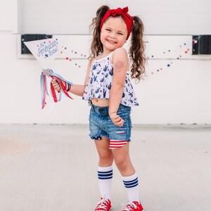 May include: A young girl wearing a red headband, a white tank top with peace signs and stars, denim shorts with a red, white, and blue design, white socks with red and blue stripes, and red sneakers. She is holding a white flag with the text "All American Babe" in blue and red lettering.