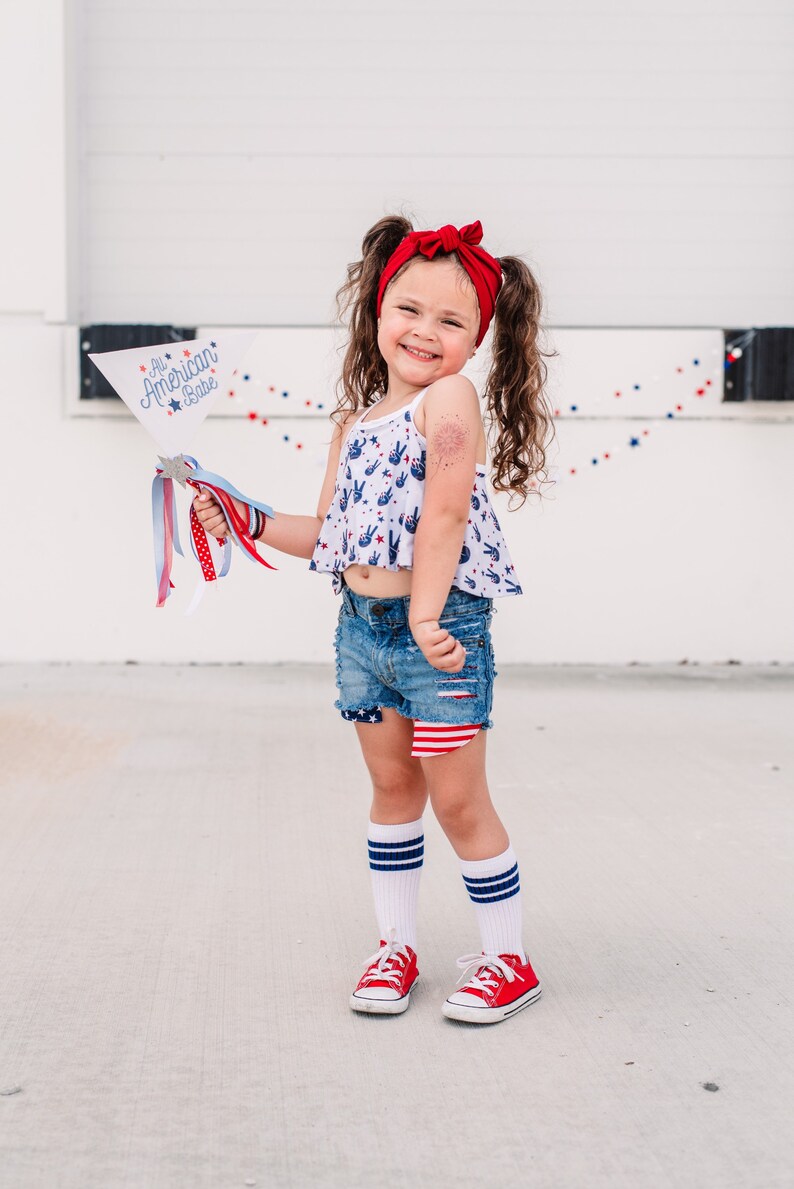 May include: A young girl wearing a red headband, a white tank top with peace signs and stars, denim shorts with a red, white, and blue design, white socks with red and blue stripes, and red sneakers. She is holding a white flag with the text "All American Babe" in blue and red lettering.