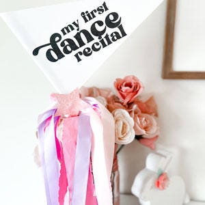 Könnte beinhalten: Ein weißes dreieckiges Banner mit dem Text "my first dance recital" in Schwarz. Das Banner ist an einer Vase mit rosa und violetten Bändern befestigt.