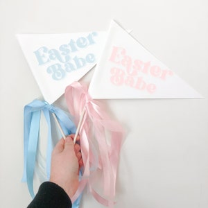 May include: Two white triangular flags with blue and pink text that reads "Easter Babe". The flags are attached to wooden sticks with blue and pink ribbons.