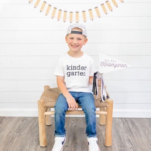 May include: A young boy wearing a white t-shirt with the text "kinder garten" printed on it, blue jeans, and a black and white patterned baseball cap. He is holding a white sign that says "1st day Kindergarten" and is sitting on a wooden stool.